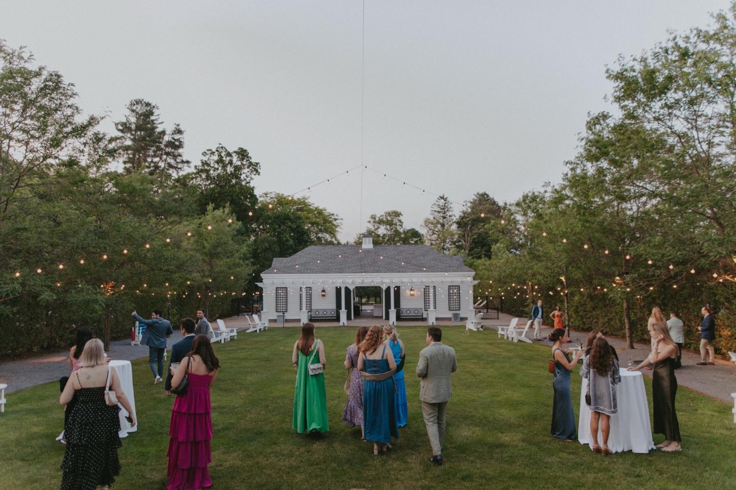 Guests in formal attire gather outside a white house with string lights.
