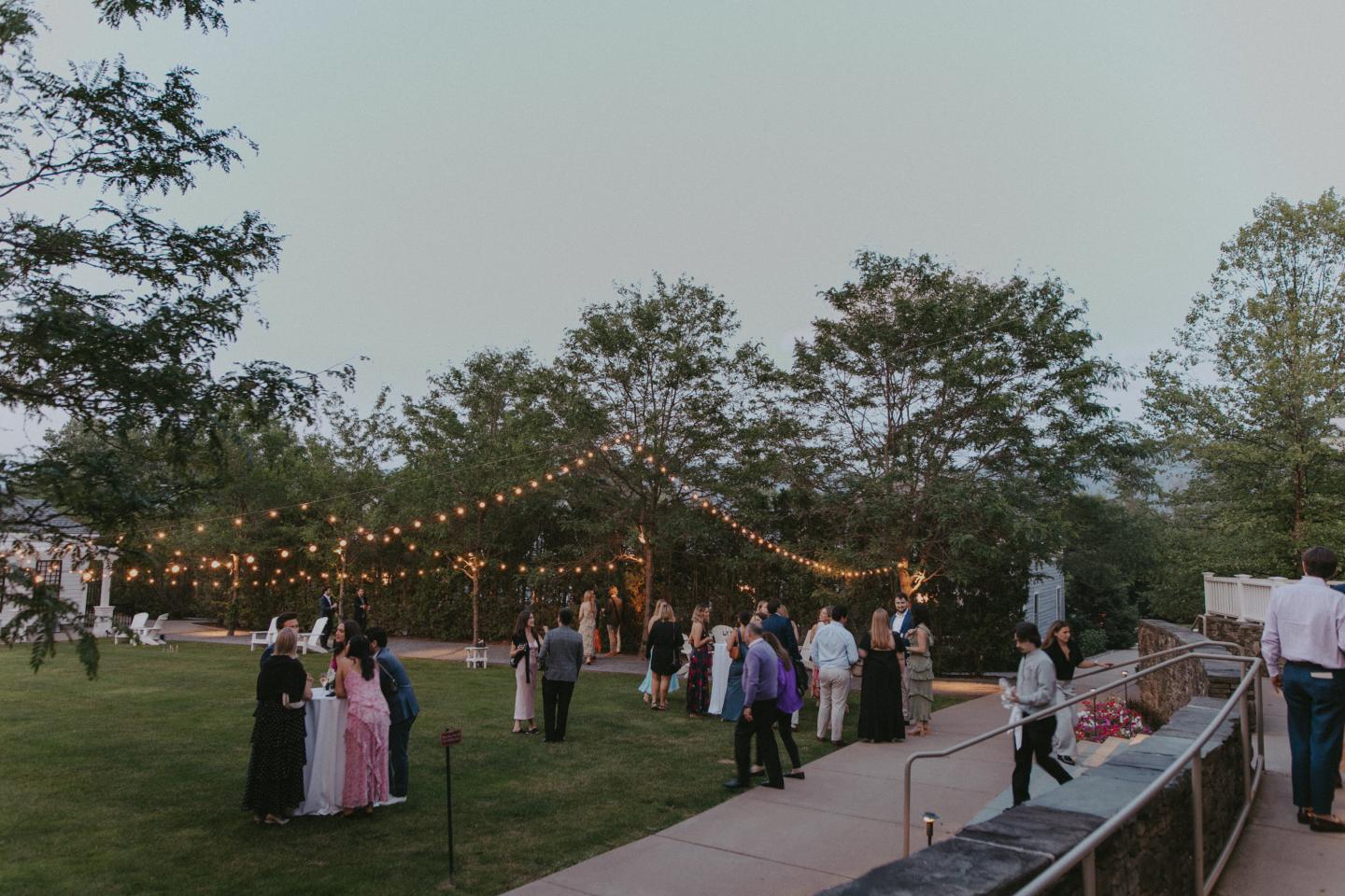 Gathering on a lawn with string lights and people in formal wear.