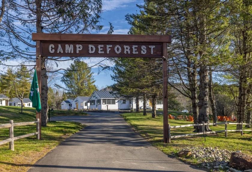 Camp entrance with sign, surrounded by trees and path leading to buildings.