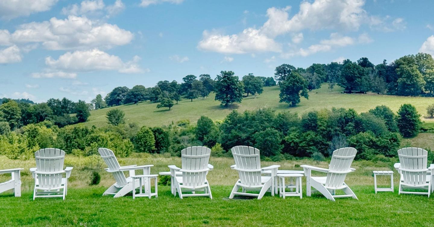 White Adirondack chairs overlooking green hills under a blue, cloudy sky.