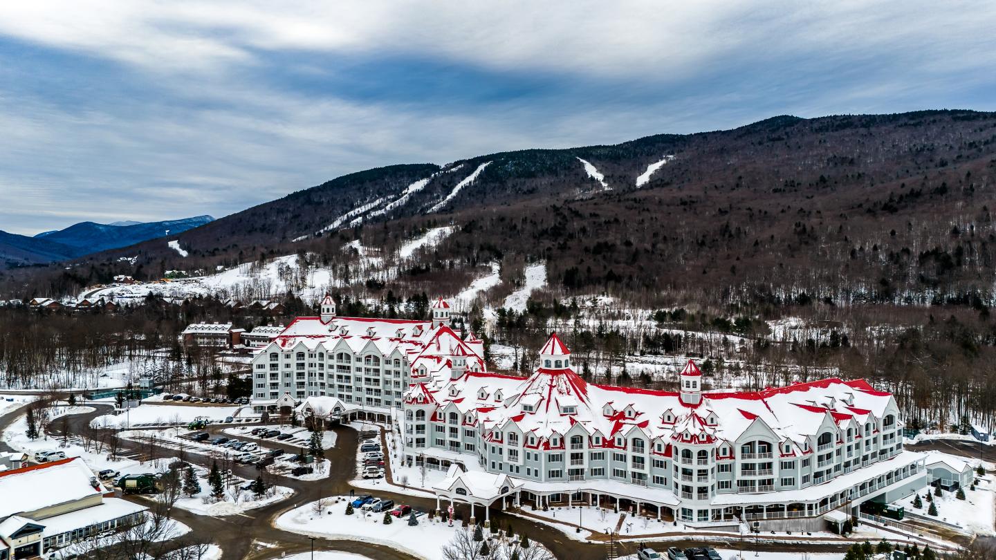 Snowy resort with red roofs set against a mountainous backdrop under a cloudy sky.