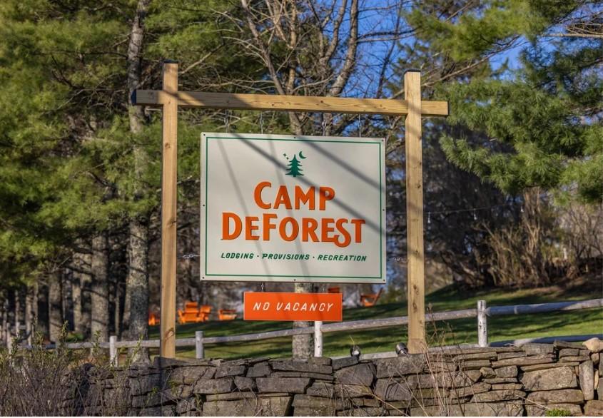Camp Deforest sign near trees and stone wall.