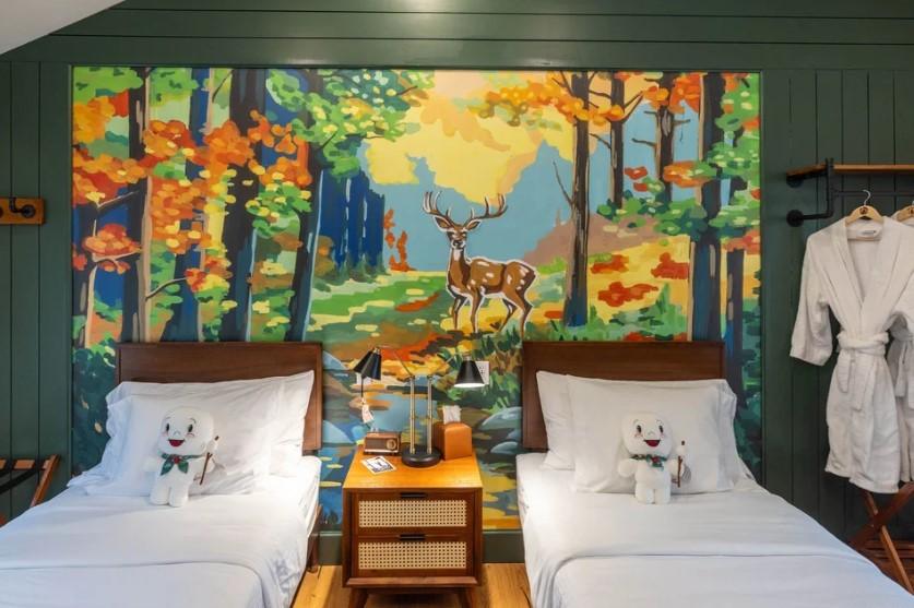 Two beds with plush toys, colorful forest mural, and robes hanging beside.