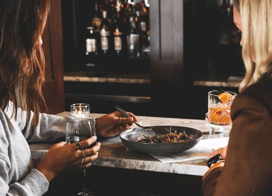 Two people dining at a bar with drinks and a bowl of food.