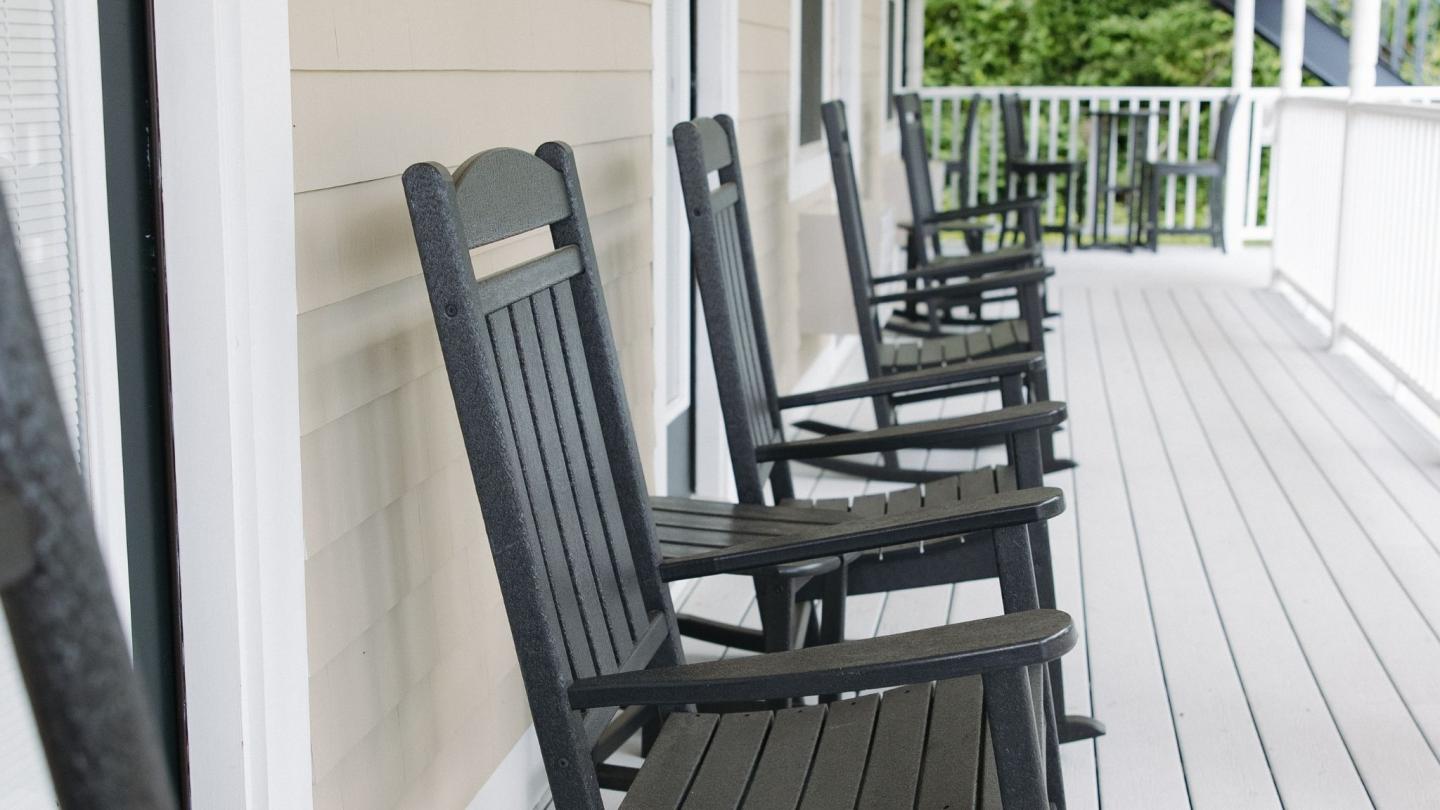 Four black rocking chairs on a wooden porch, with a green garden view.
