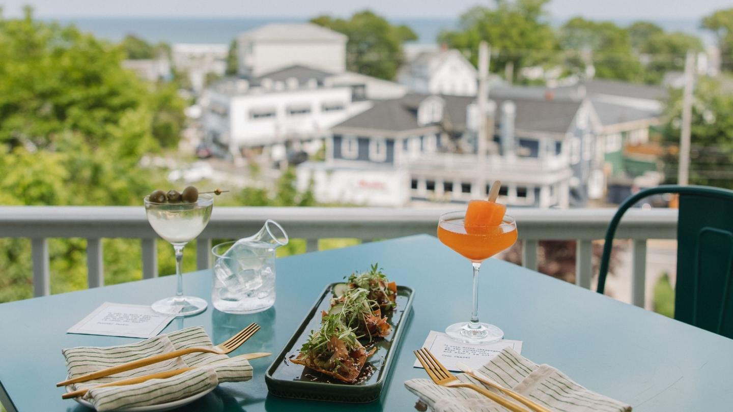 Outdoor dining table with cocktails and appetizers, overlooking buildings and trees.