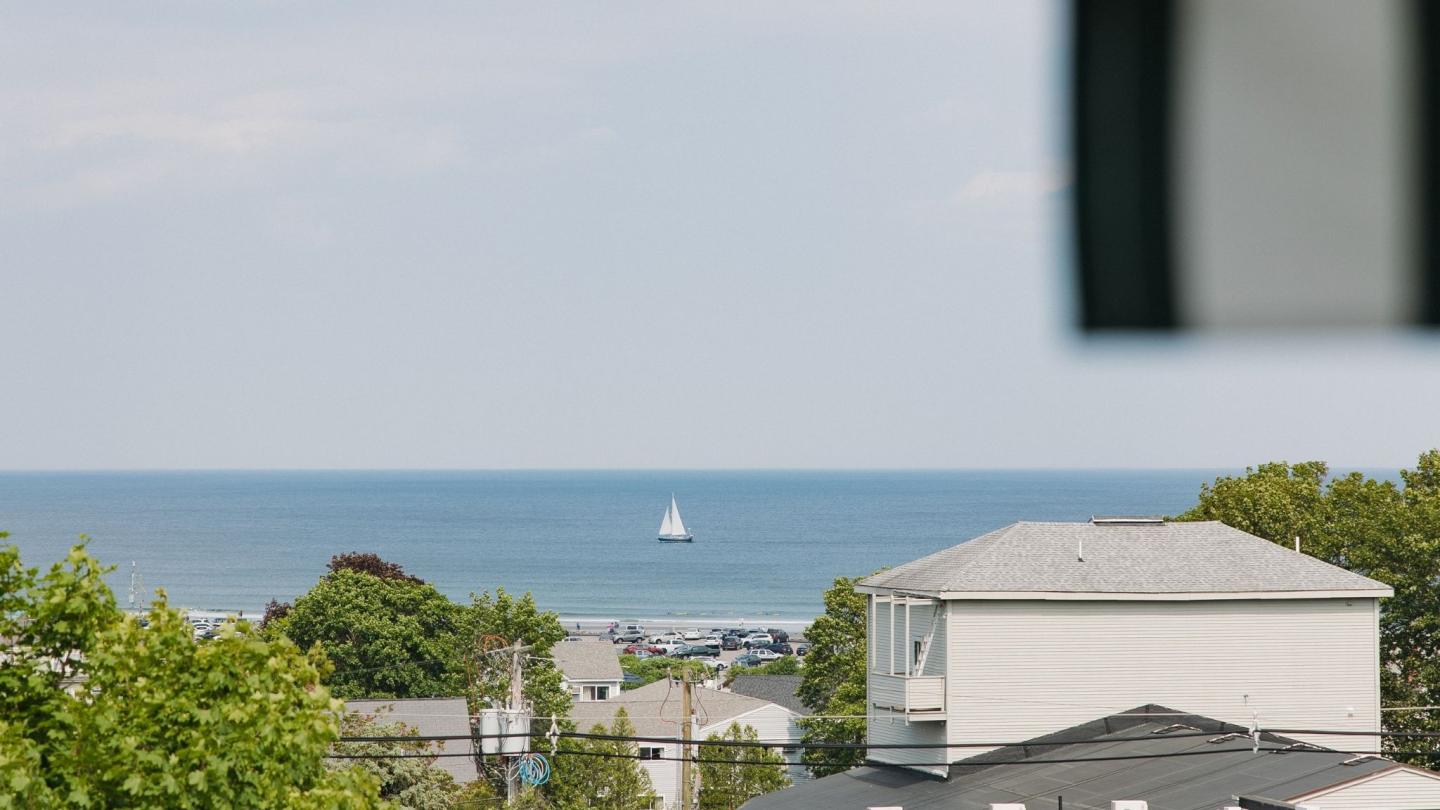 View of a sailboat on the ocean, seen over rooftops and greenery.