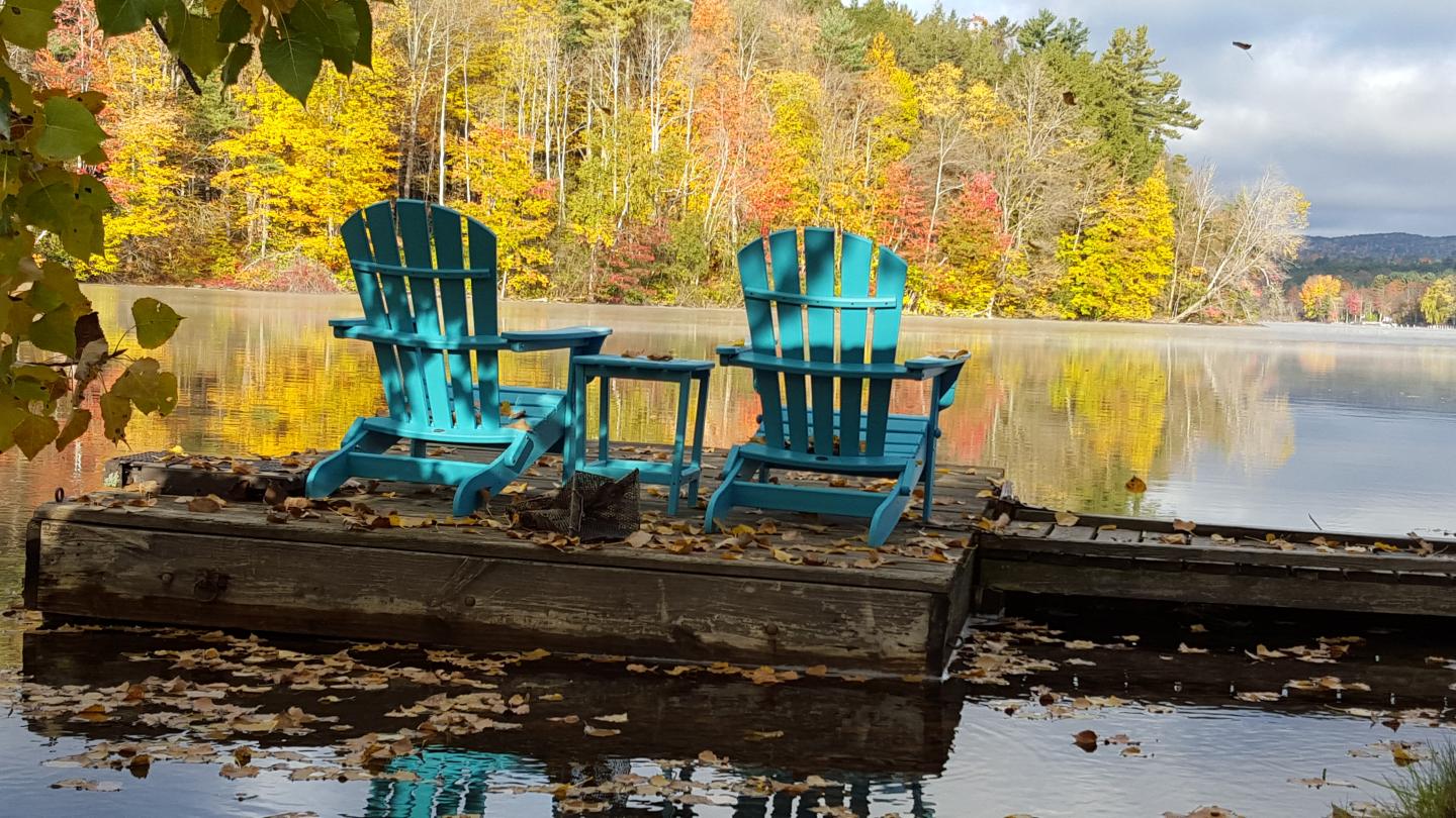 Two blue chairs on a lakeside dock, surrounded by autumn trees.