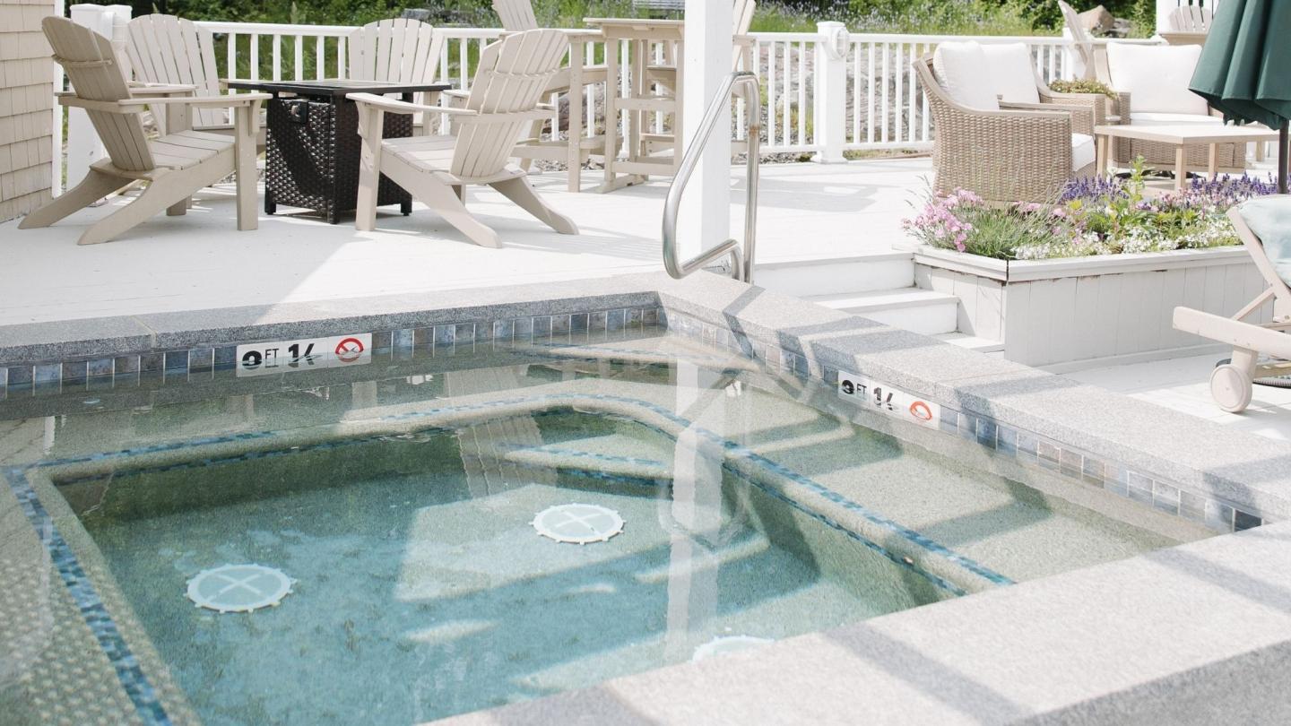 Hot tub on a sunlit patio with chairs and potted flowers nearby.