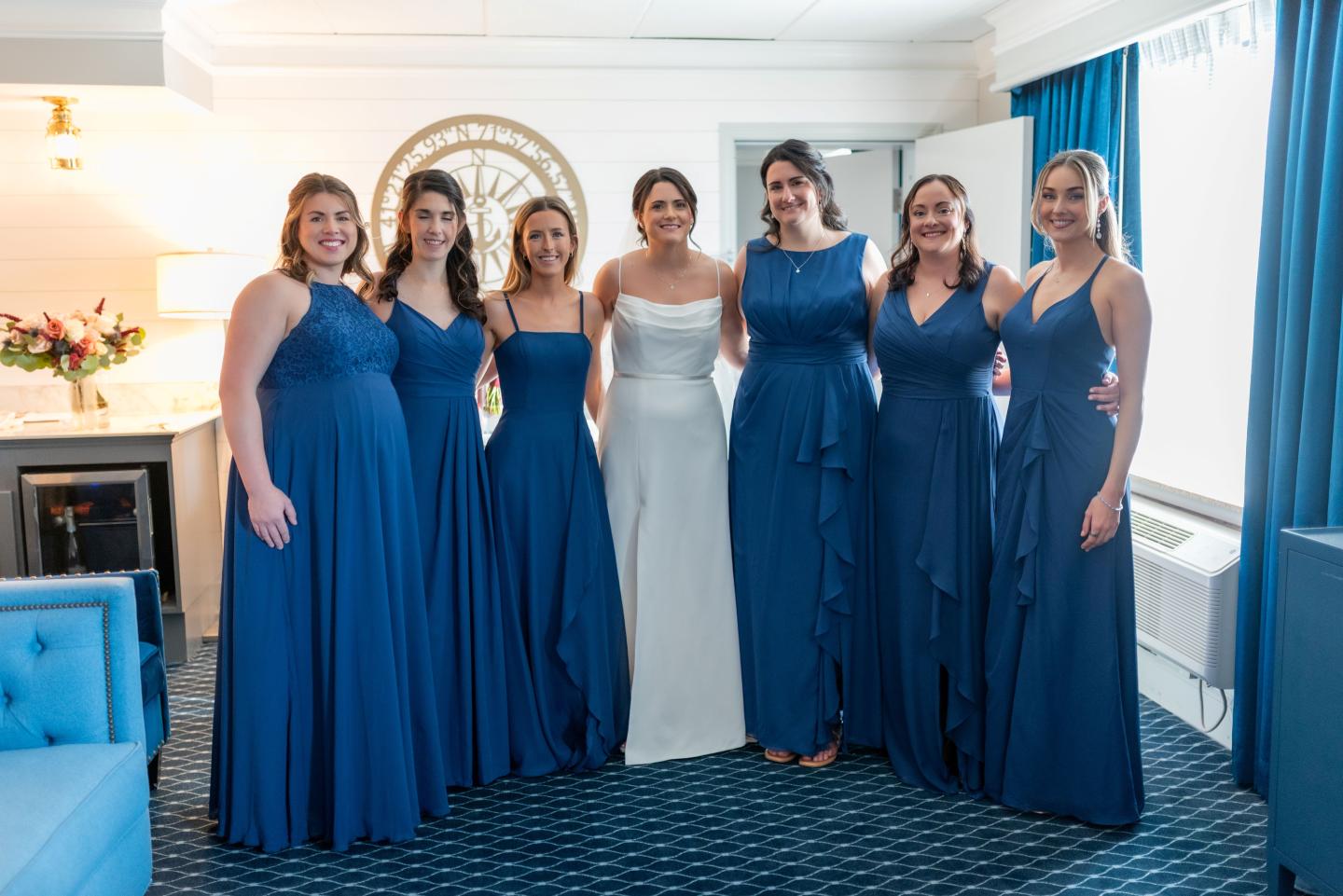 Bridal party with bride in white and bridesmaids in blue dresses, smiling in a bright room.