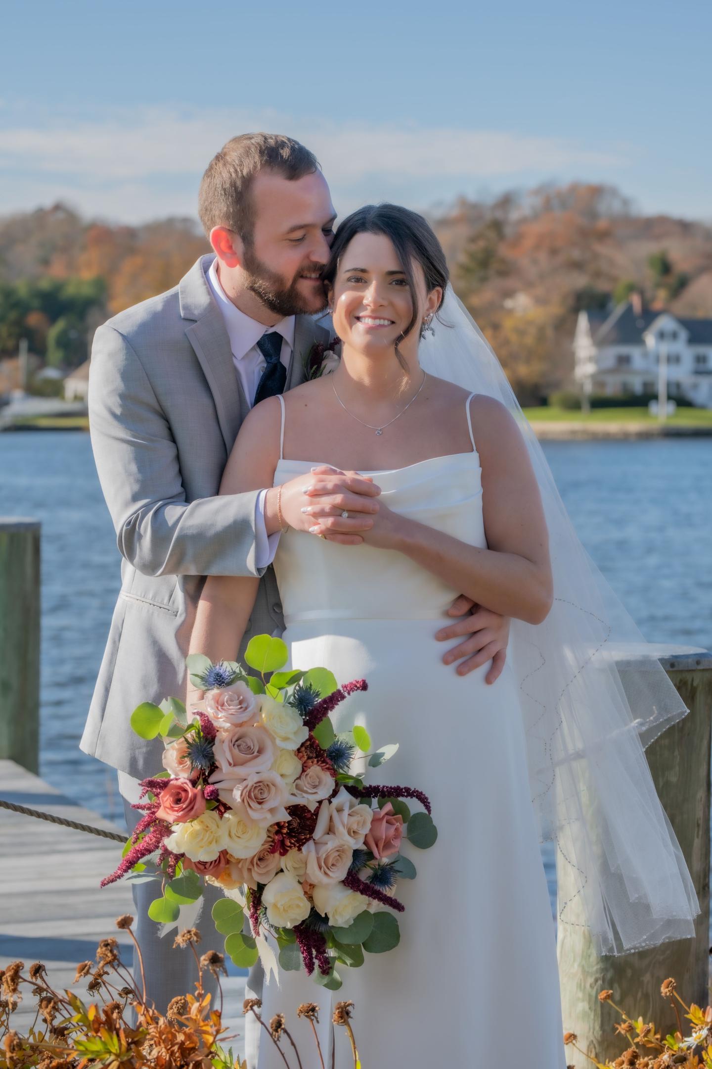 Bride and groom smiling by a lakeside, holding a floral bouquet.