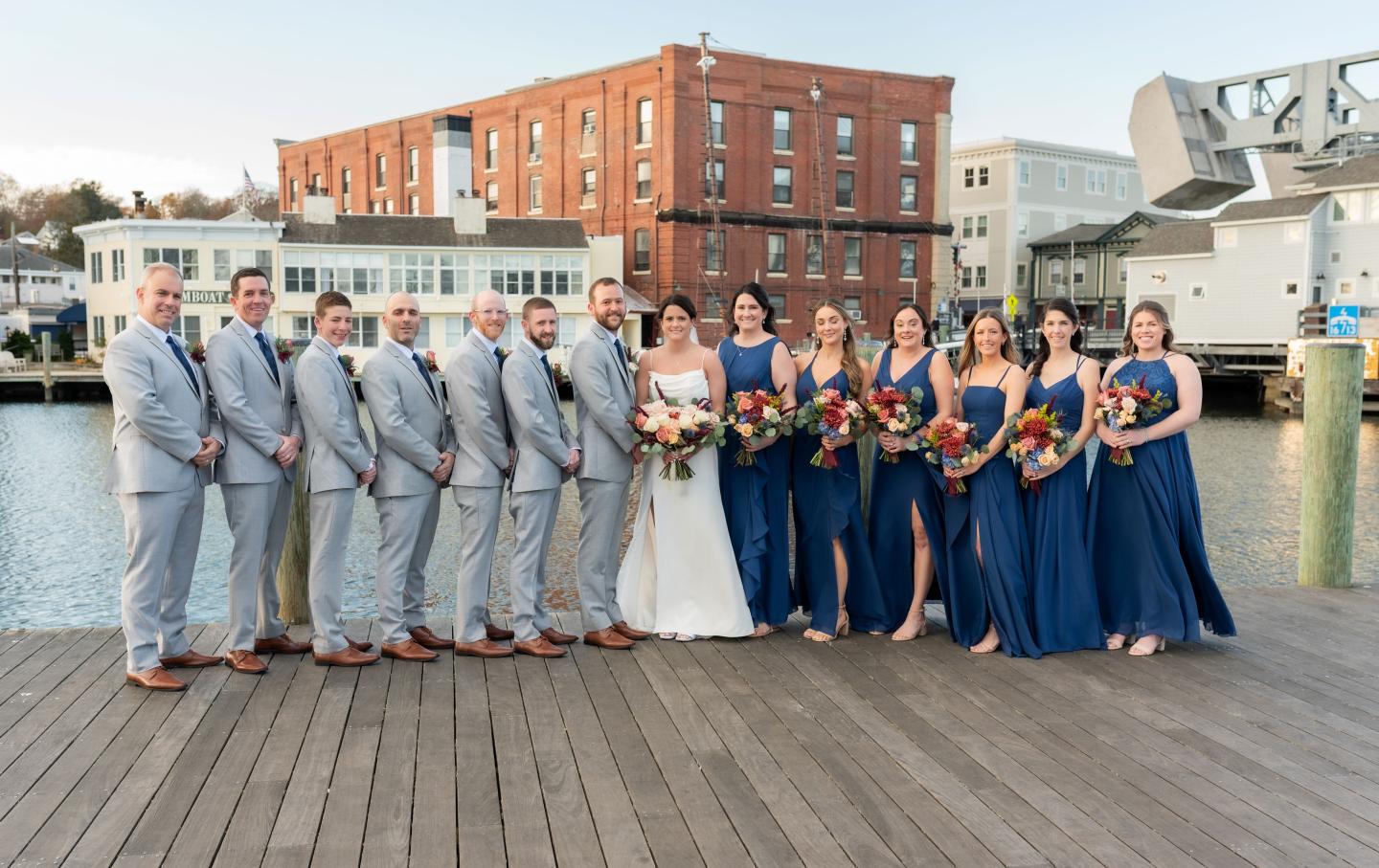 Wedding party posing on a dock, bridesmaids in blue dresses, groomsmen in grey suits.