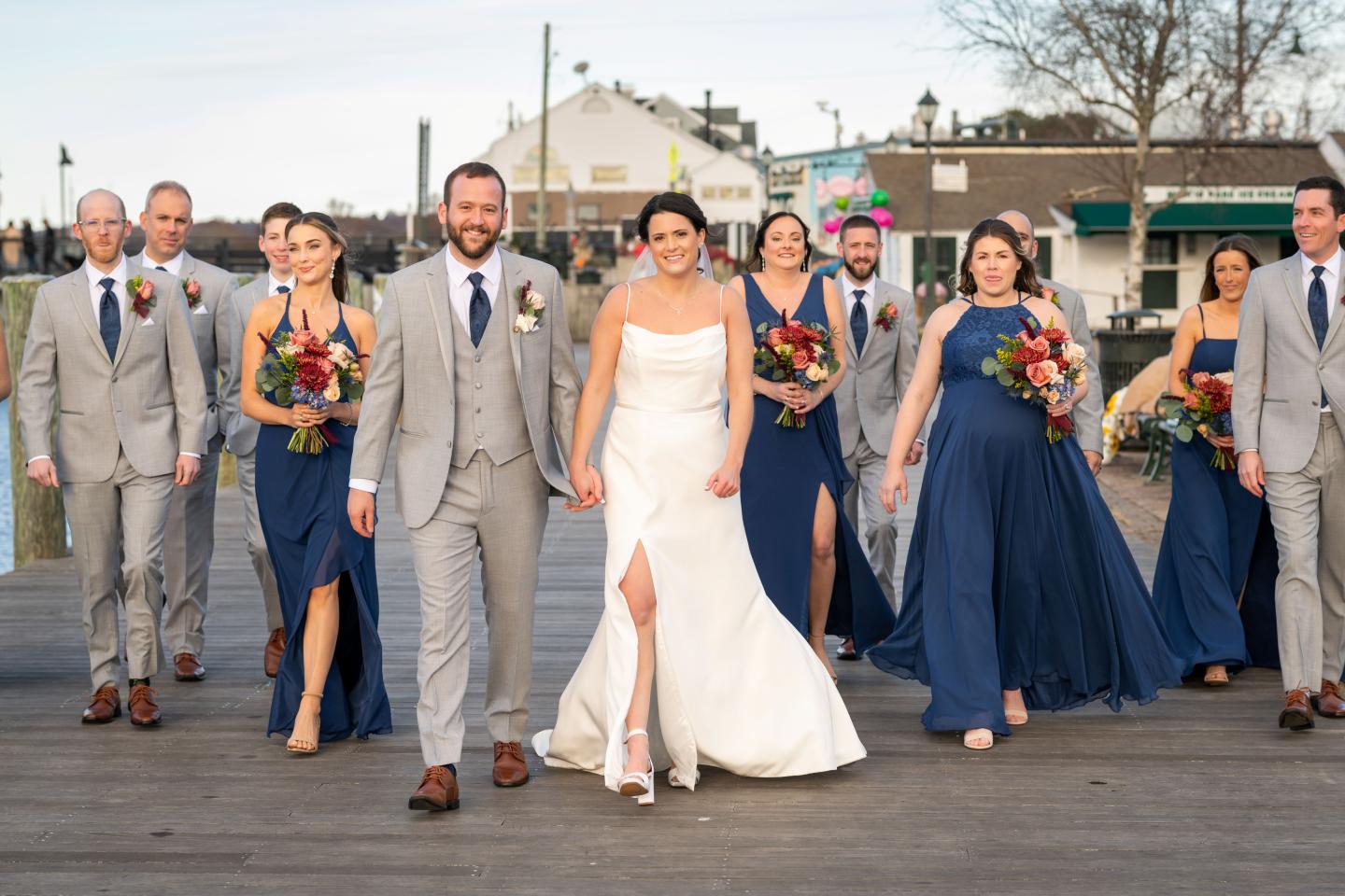 Wedding party walking on a dock, bride in white, bridesmaids in blue, groomsmen in gray.