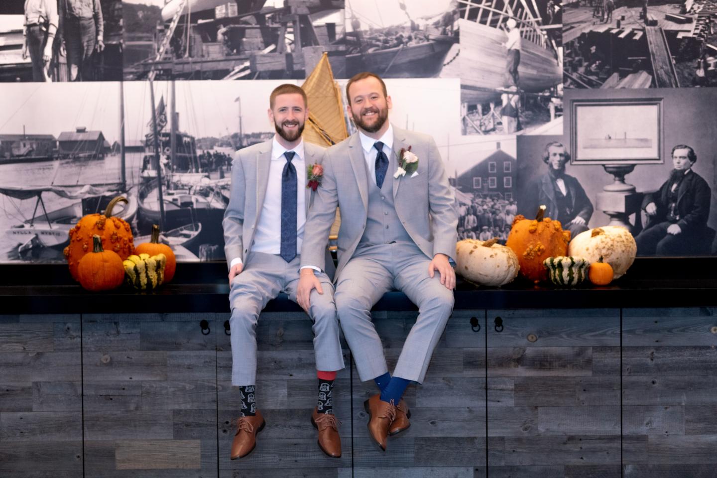 Two smiling men in suits sitting on a counter with pumpkins, in a room with vintage photos.