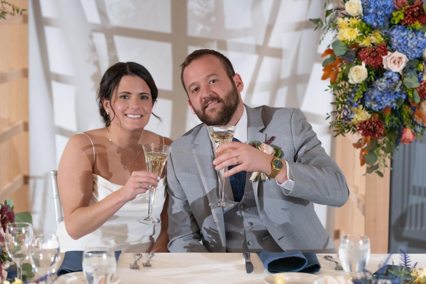 Bride and groom smiling, holding champagne glasses at a decorated table.