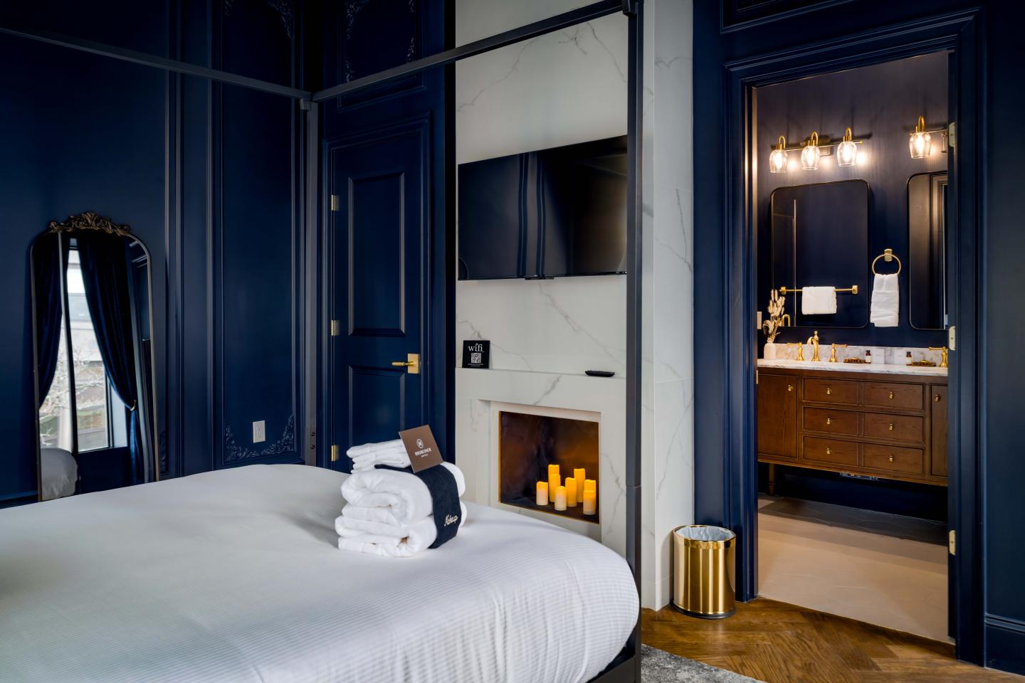 Elegant bedroom with navy walls, a bed, fireplace, and view into a lit bathroom.