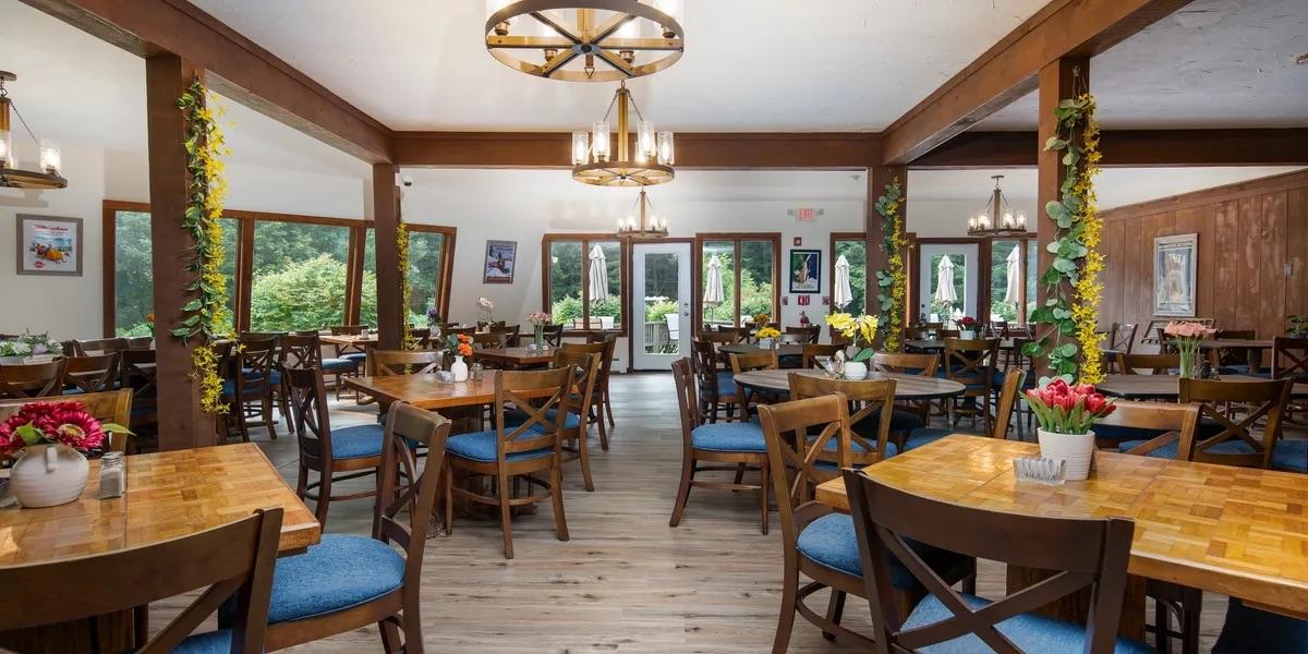 Cozy restaurant with wooden tables, blue chairs, and flower vases. Bright natural light.
