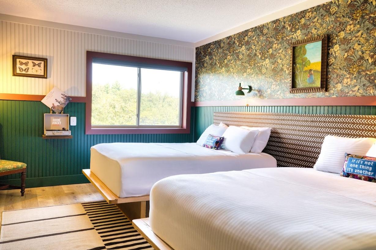 Two neatly made beds in a cozy room with patterned wallpaper and a window view.