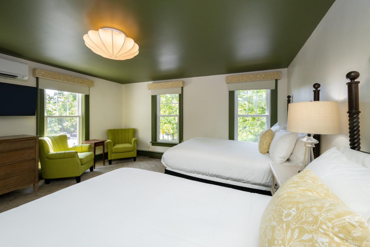 Bright hotel room with two beds, green ceiling, armchairs, and soft lighting.
