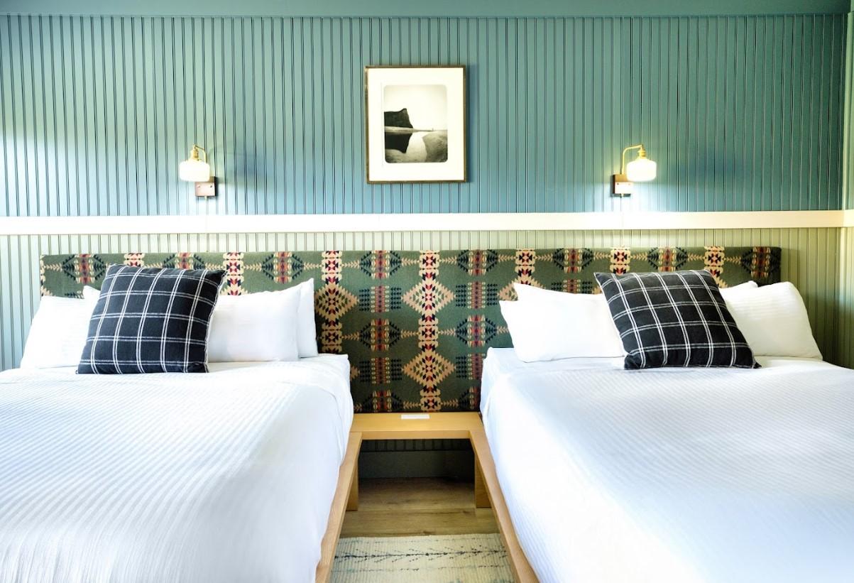 Two beds with black plaid pillows in a teal-painted room with a framed photo.