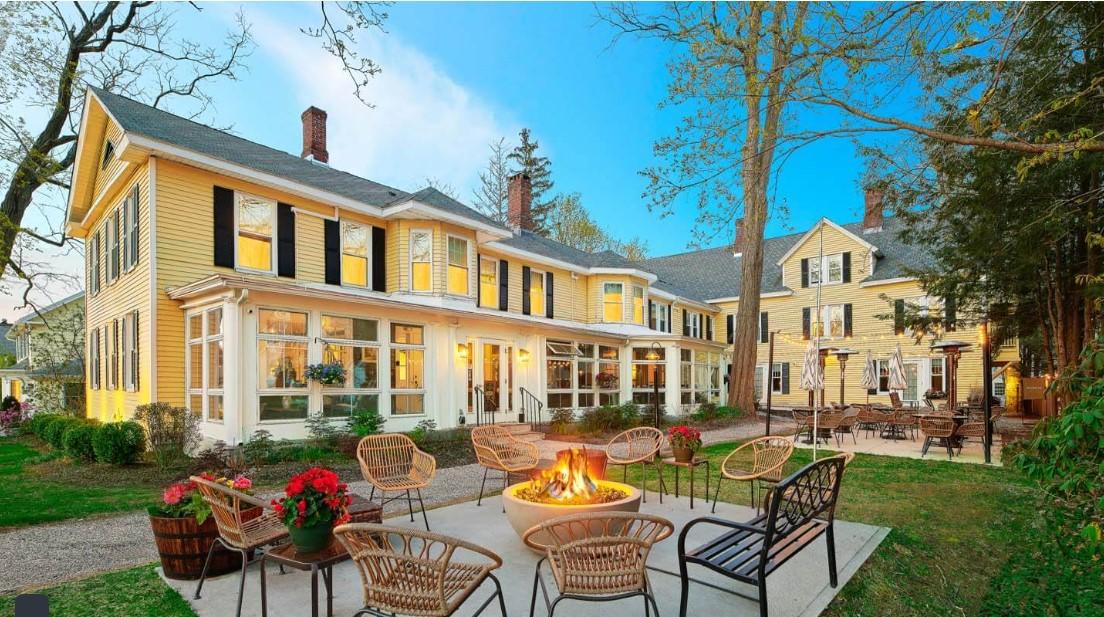 Yellow house with a patio, fire pit, and cozy seating, surrounded by trees.