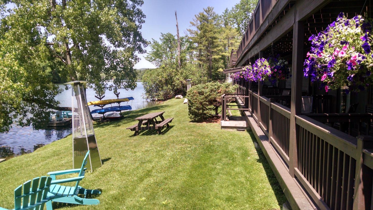 Lakeside lawn with chairs, picnic table, and flower-adorned building. Canoes by water.