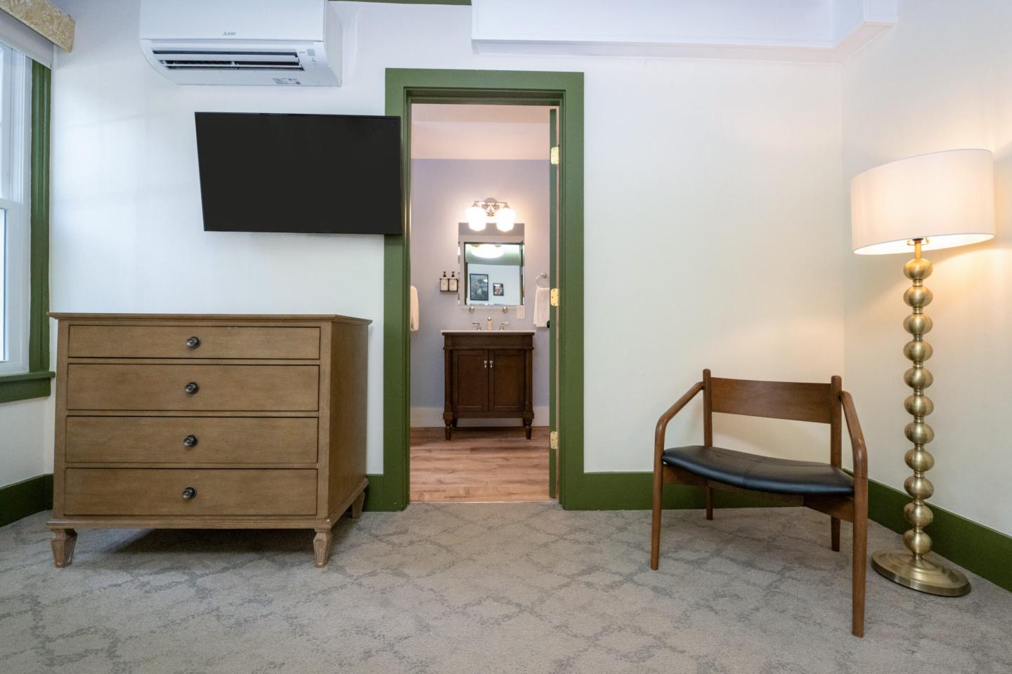 Cozy room with a dresser, wall-mounted TV, chair, and a view into a bathroom.
