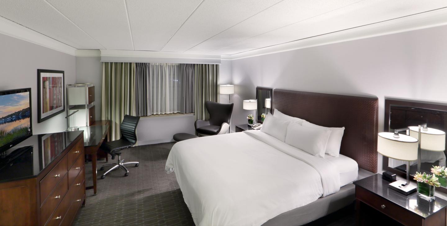 Hotel room with a large bed, desk, chairs, and a TV. Neutral-toned decor.