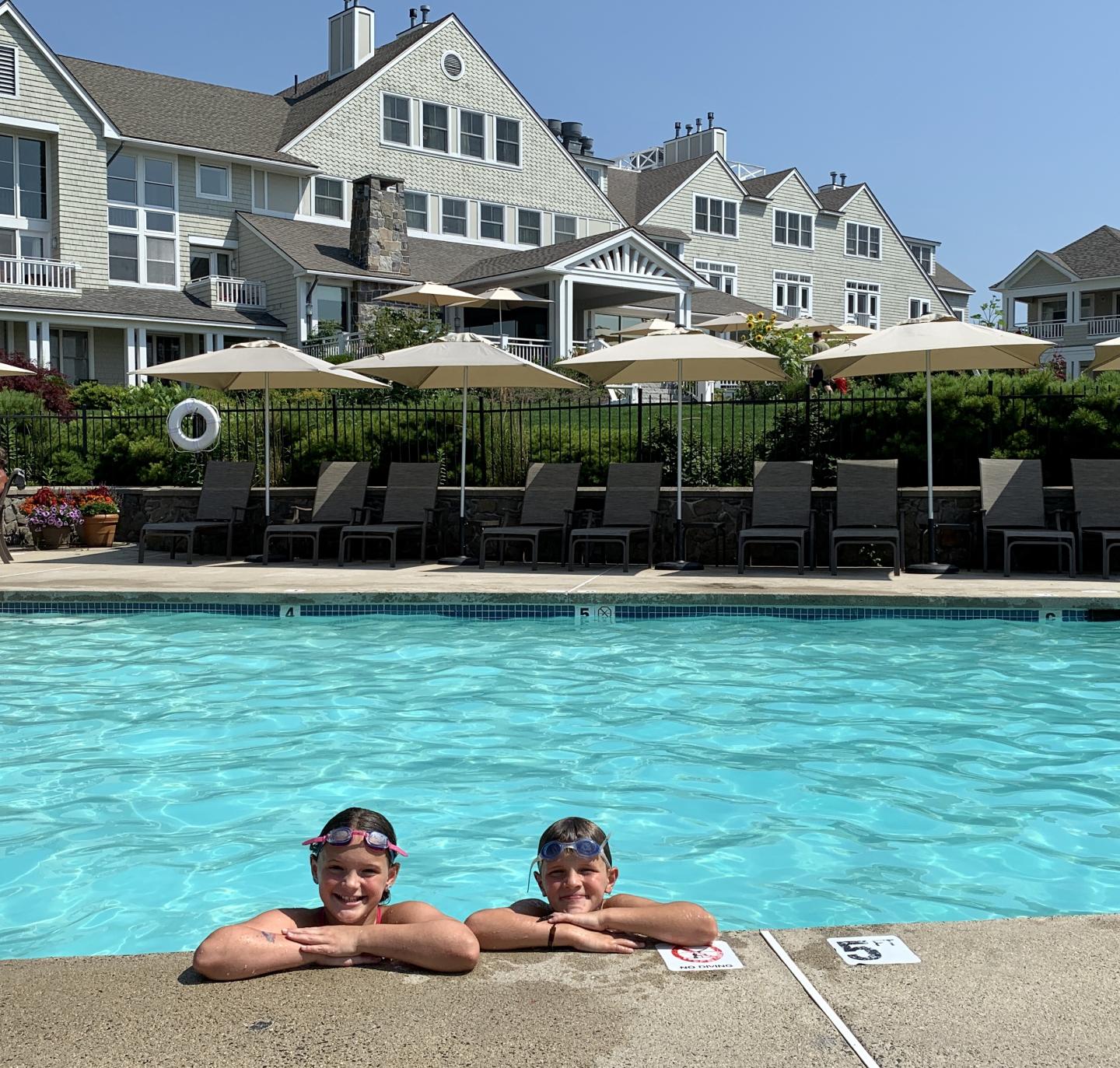 Two kids in a swimming pool, smiling with houses in the background.