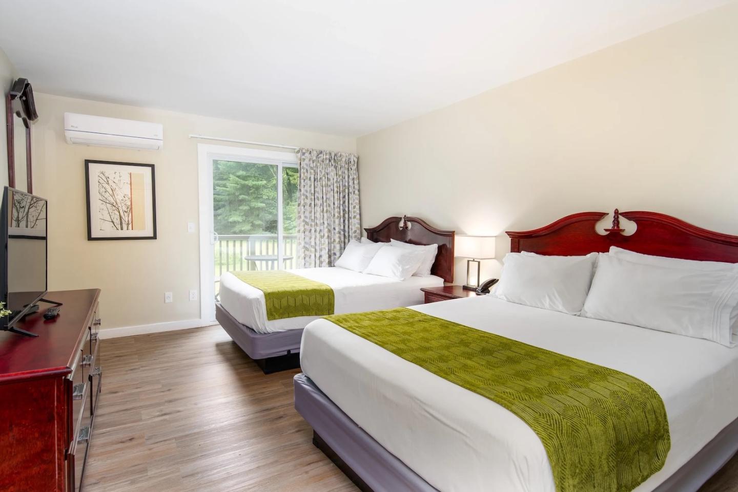 Two beds in a hotel room with green throws, wooden furniture, and a view outside.