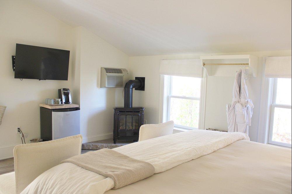 White bedroom with a bed, TV, mini fridge, and a fireplace. Bright natural light through windows.