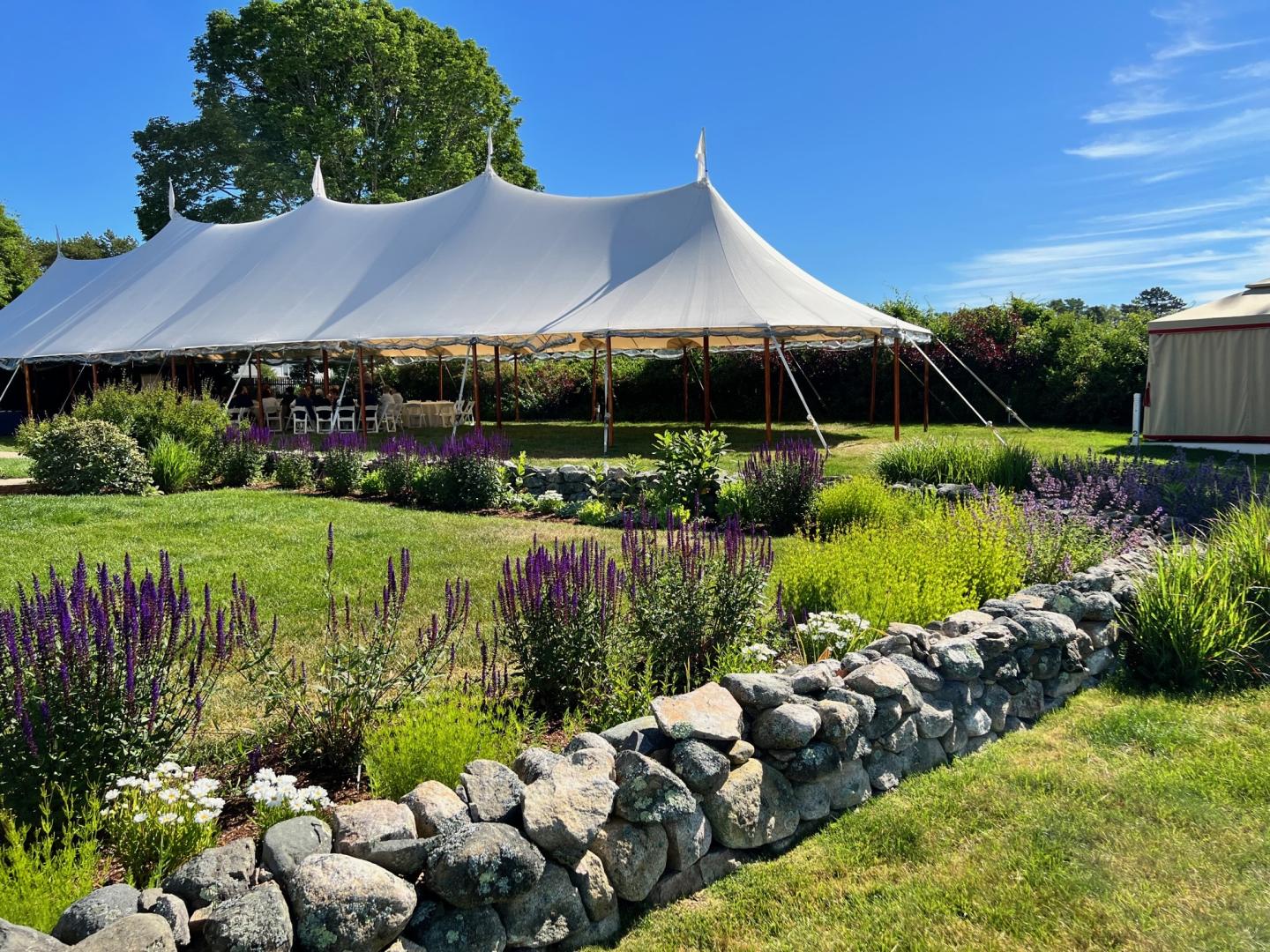 White tent on green lawn, stone wall, vibrant flowers, and clear blue sky.