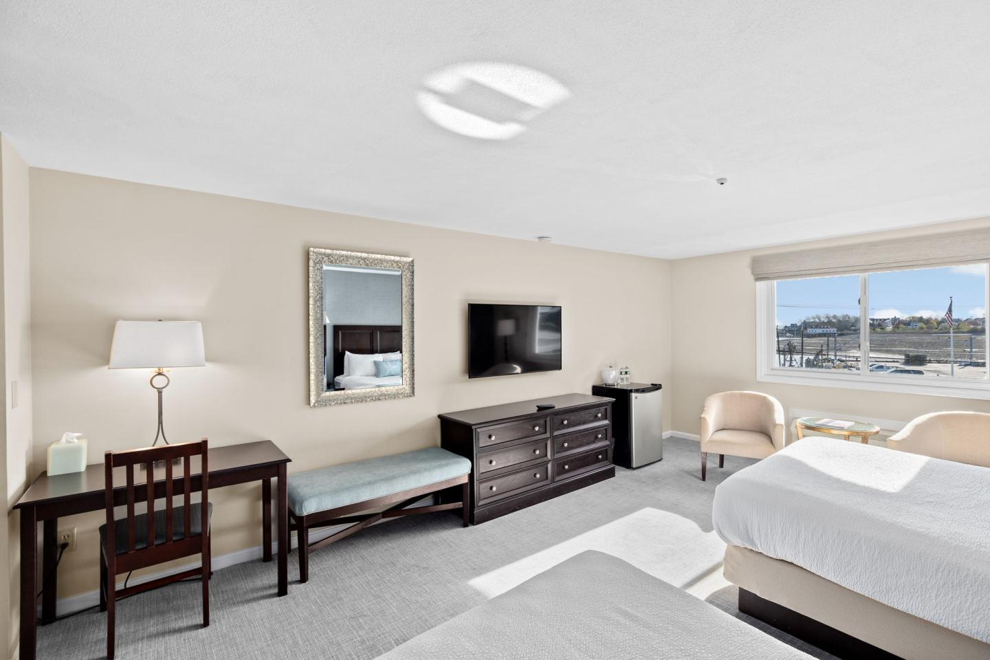 Bright hotel room with two beds, desk, TV, and harbor view from window.