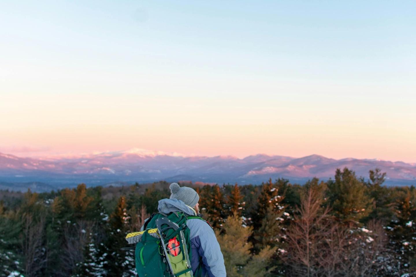 Hiker with backpack overlooks snow-covered forest and mountains at sunrise.