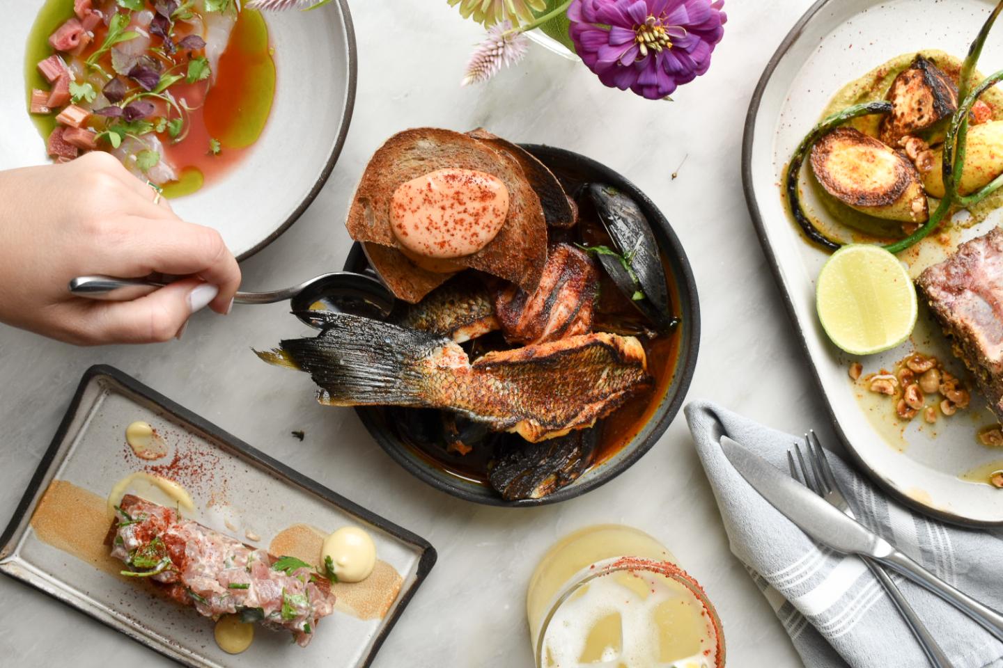Hand reaching for grilled seafood platter, surrounded by colorful dishes and flowers.