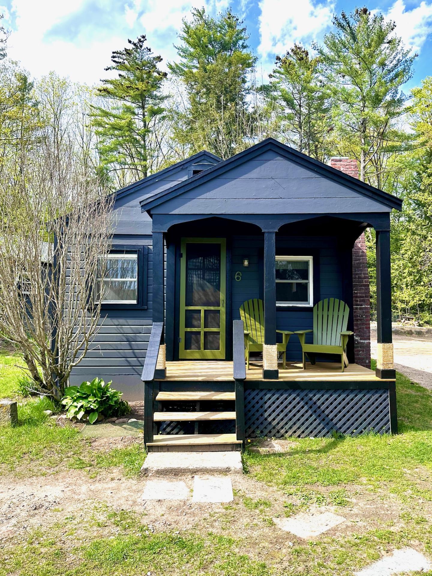 Small blue cabin with porch, green chairs, surrounded by trees.