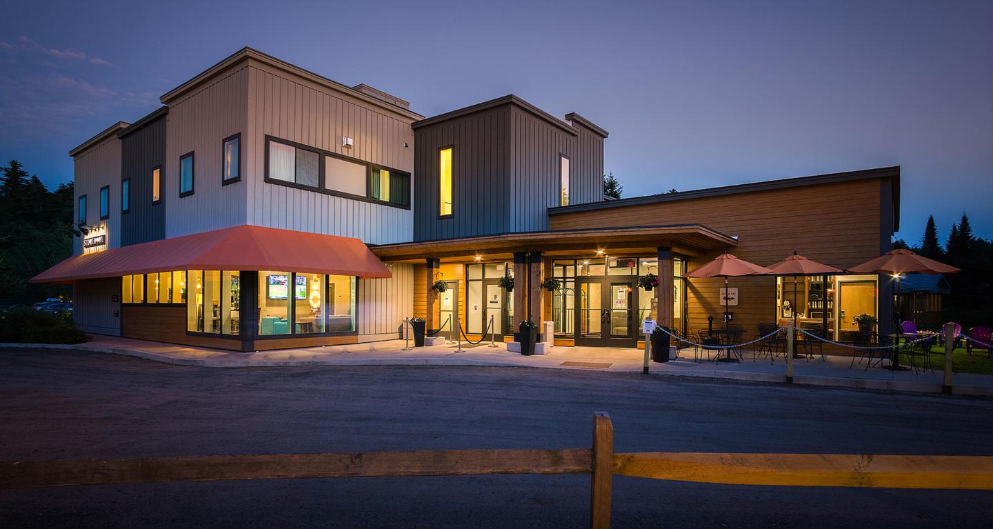 Modern cafe building at dusk, warm lights, patio umbrellas outside.