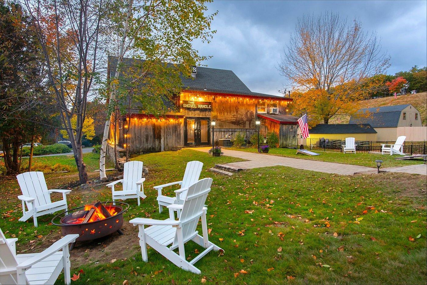 Barn with string lights, surrounded by trees and white chairs near a fire pit.