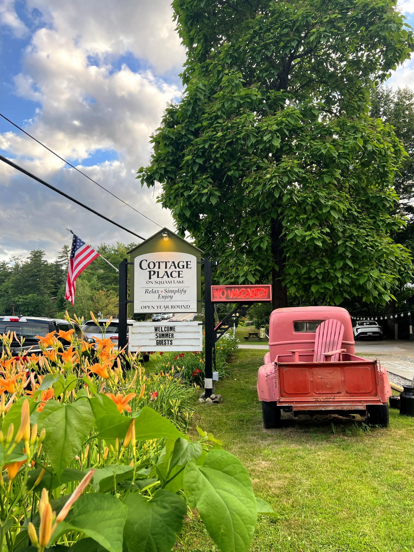 Red truck on grass beside a sign and flowers, with trees and an American flag.