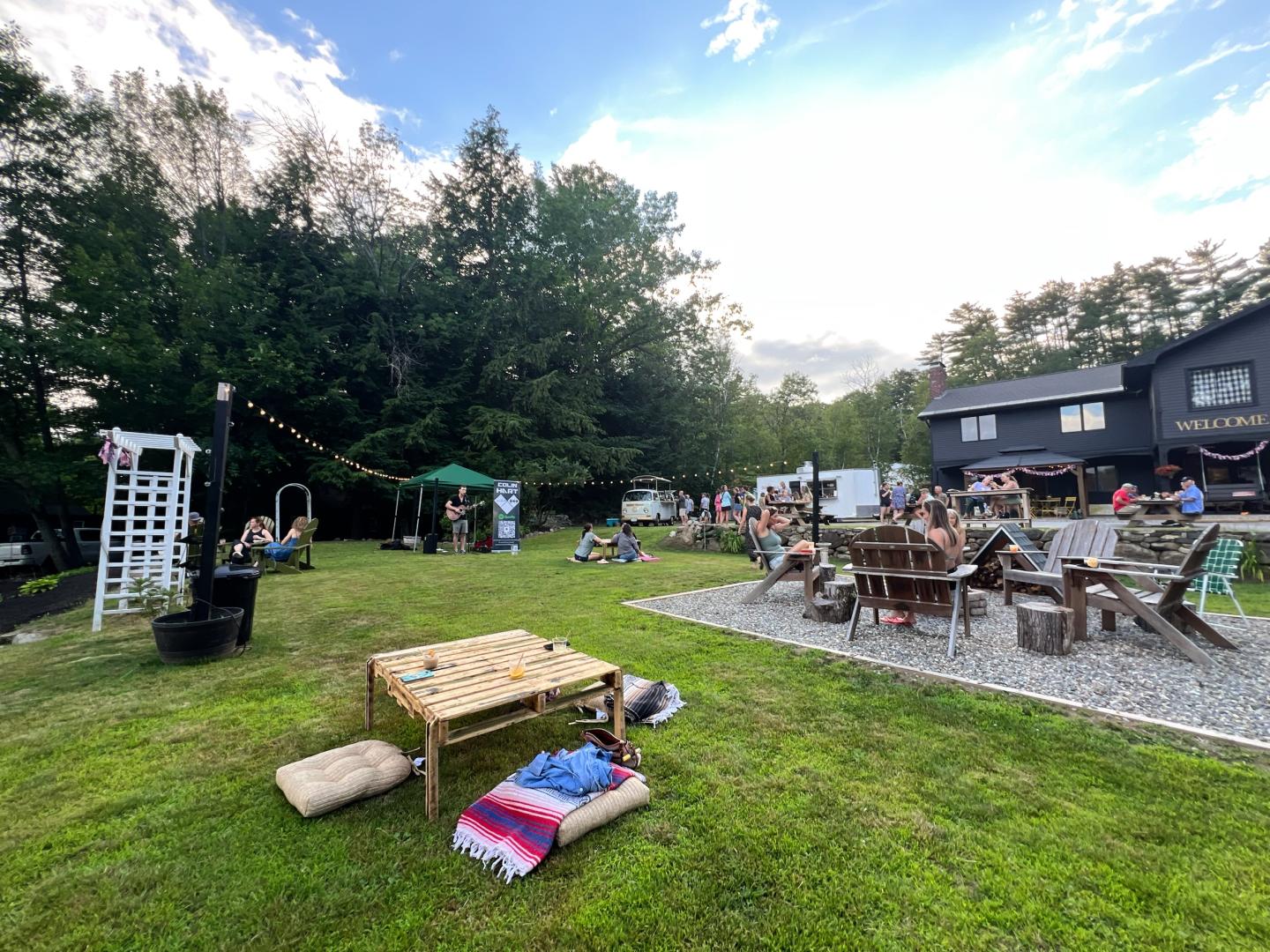 Outdoor gathering with tables on a lawn, surrounded by trees and a building.