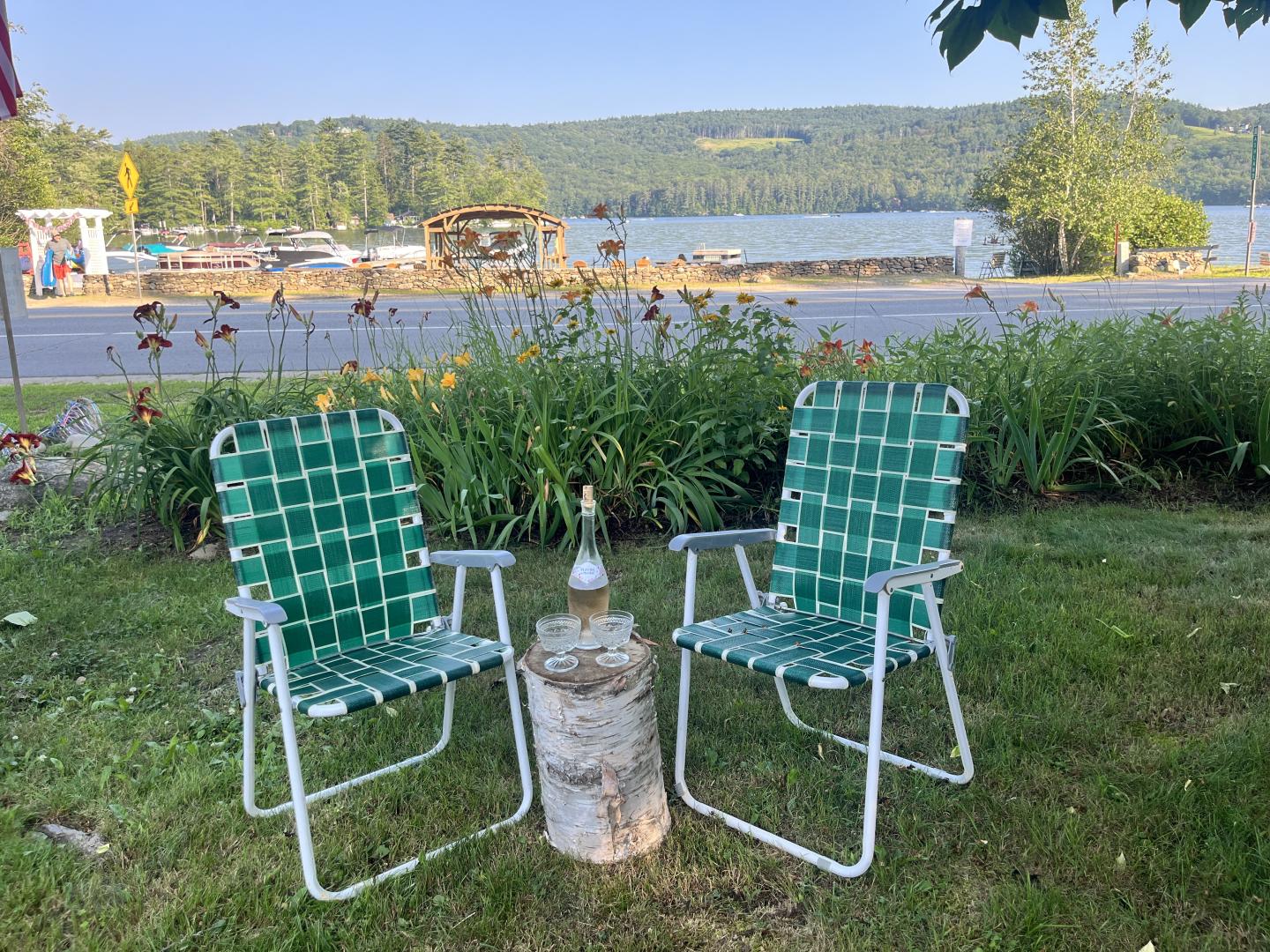 Two green lawn chairs by a lake, with a tree stump table between them.
