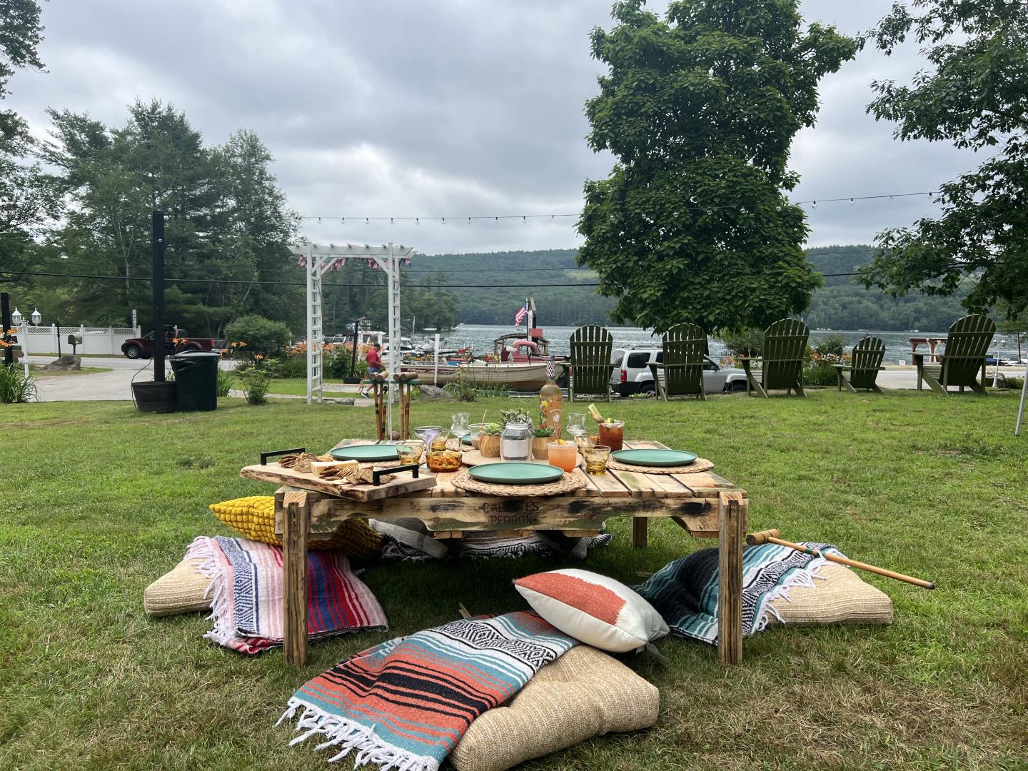Low wooden table with food, surrounded by colorful cushions on grass, scenic lake view behind.