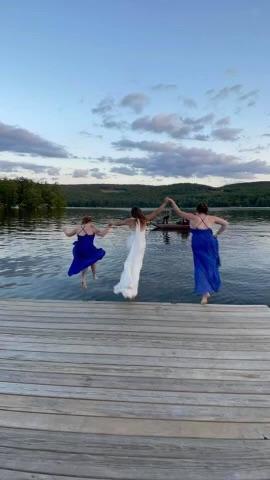 Three women in dresses jump off a dock into a lake at sunset.