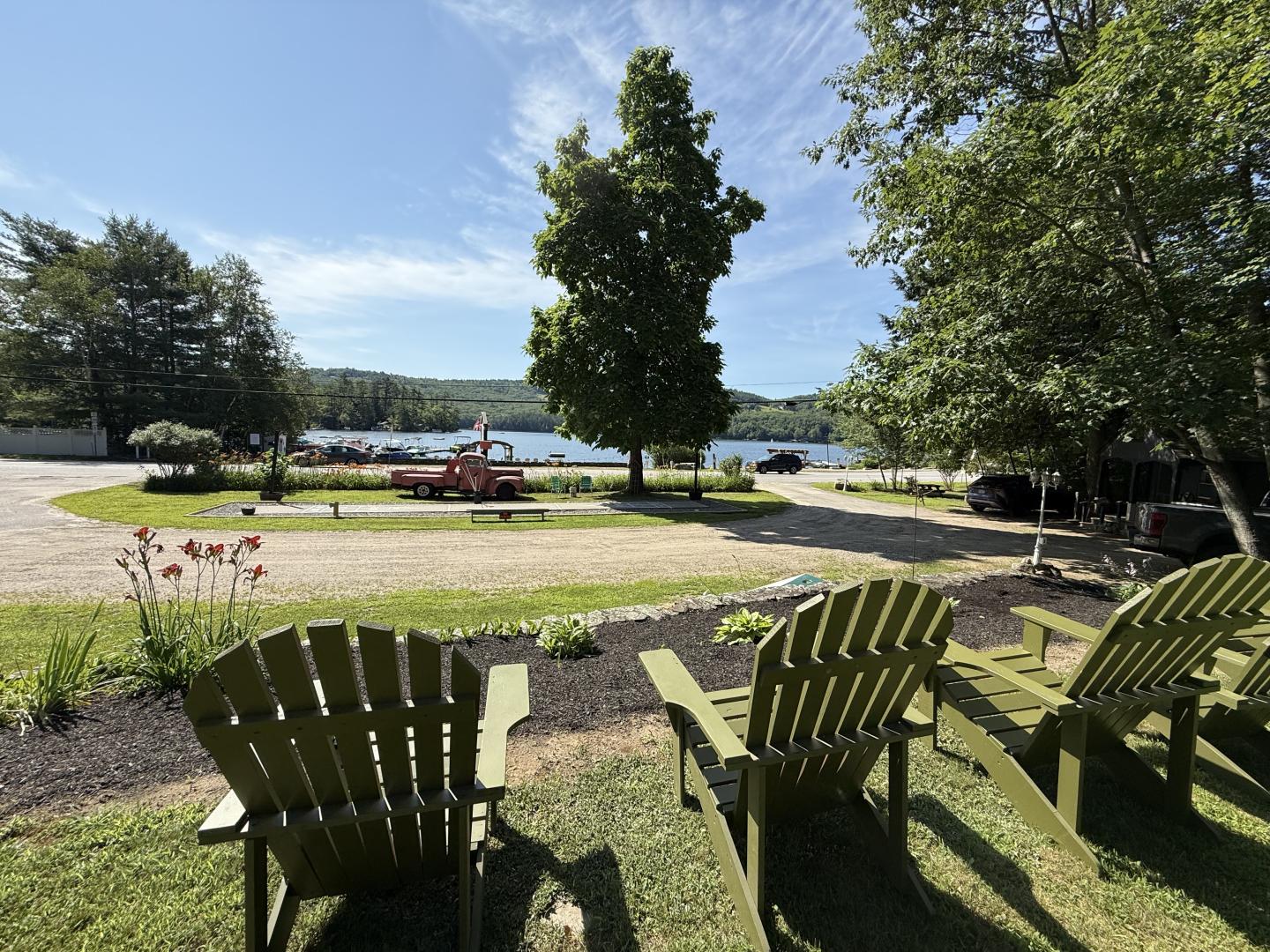 Green chairs facing a lake under a blue sky, surrounded by trees and parked cars.