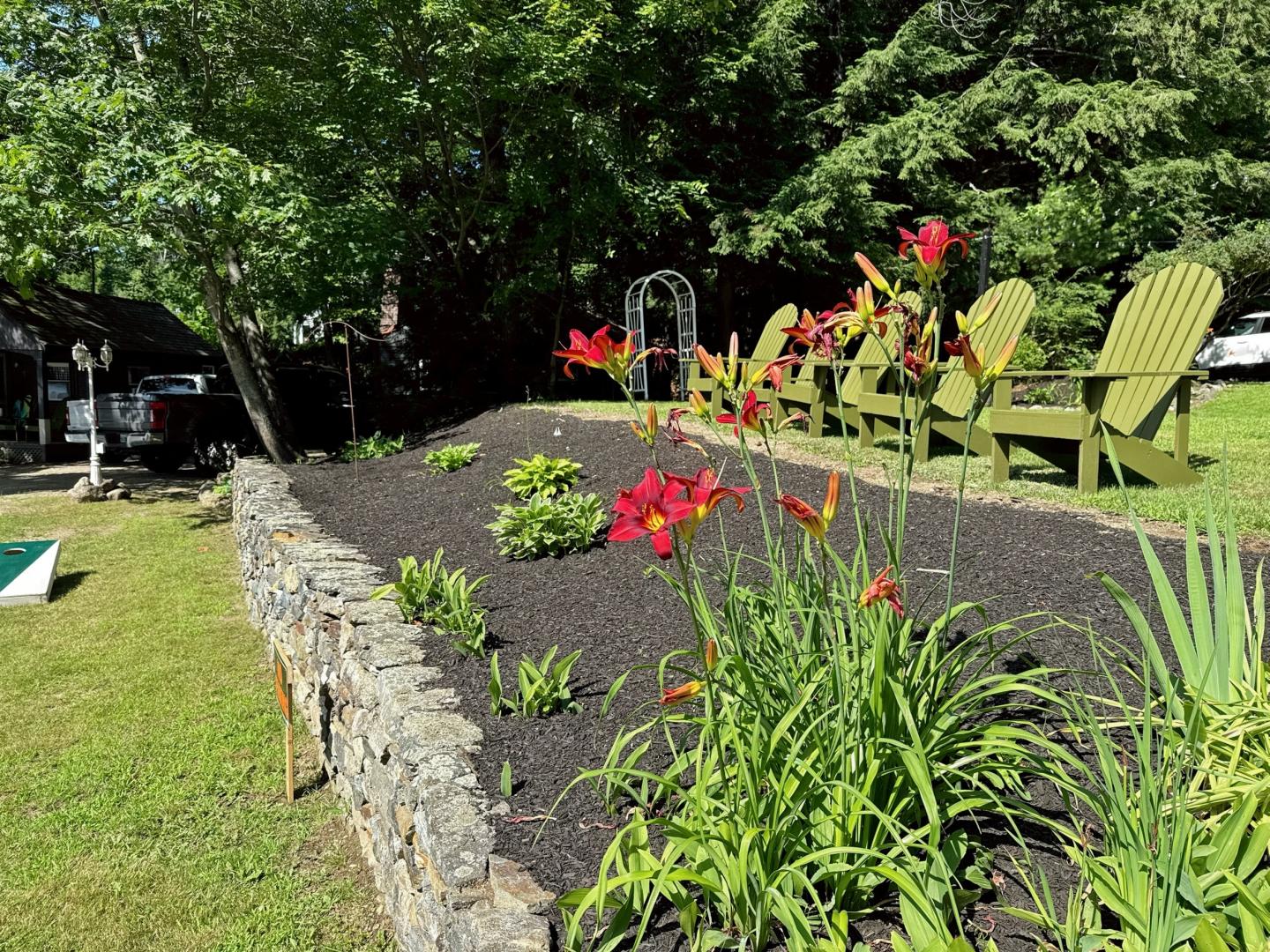 Garden with red flowers, stone border, and green chairs in sunlight.