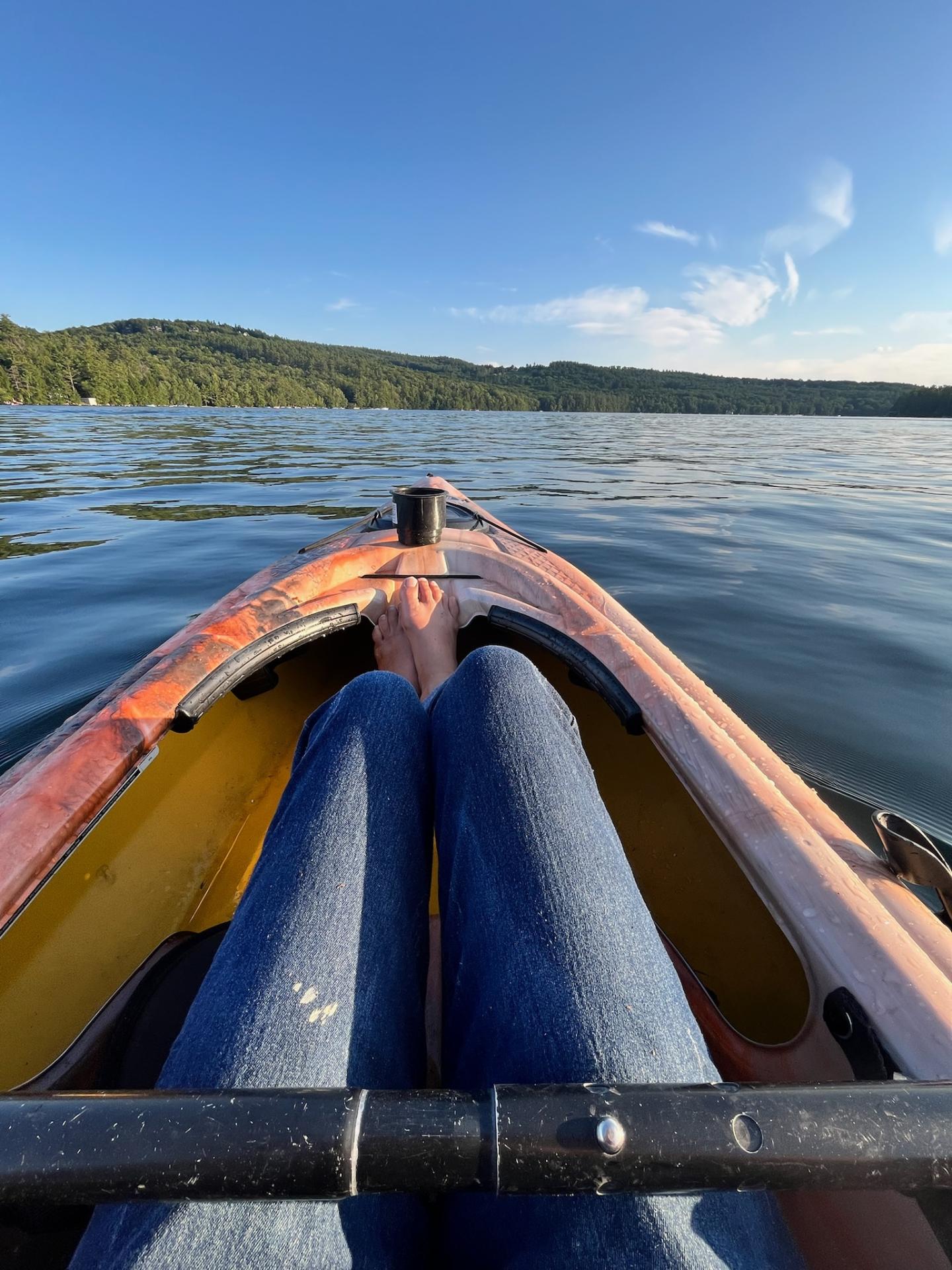 Kayak on a lake, feet inside, sunny day with distant forested shore.