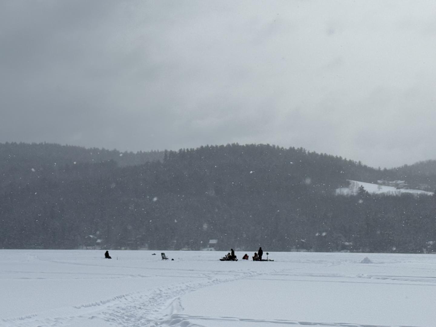 Snowy landscape with distant figures on a frozen lake, mountains in the background.
