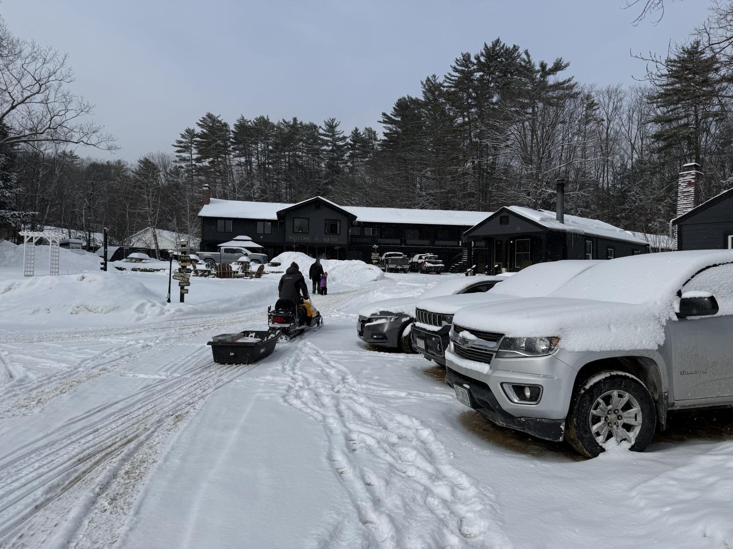 Snowy parking lot with cars and sleds, cabins and trees in the background.