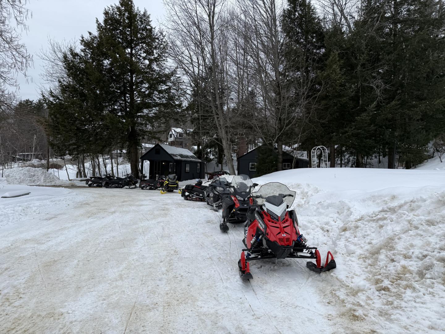 Snowmobiles parked on a snowy path, surrounded by trees and small cabins.
