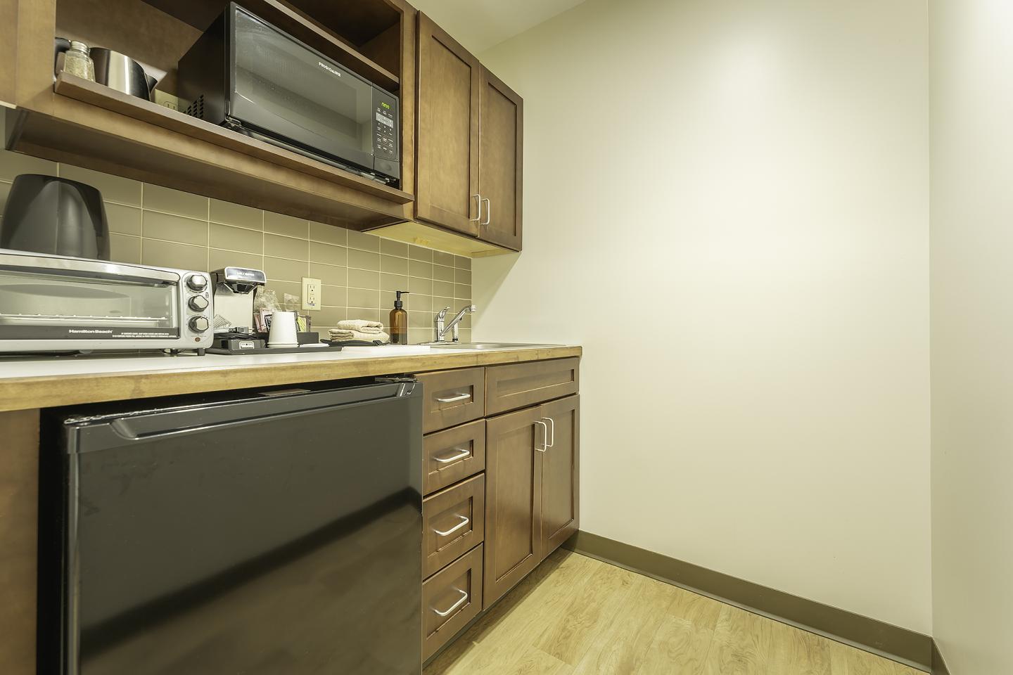 Compact kitchenette with wood cabinets, microwave, mini-fridge, and a tiled backsplash.